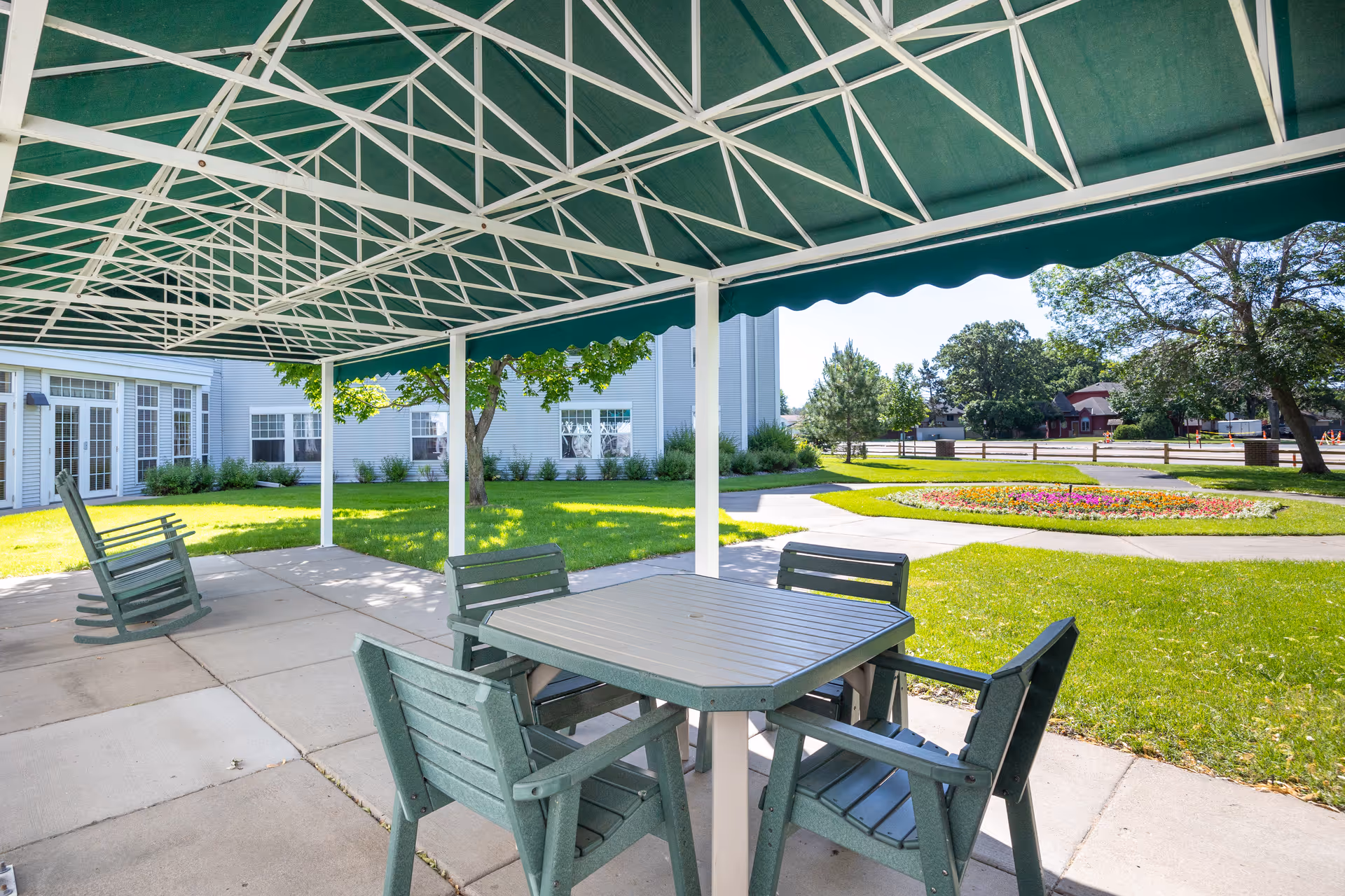 Outdoor covered patio area with a green canopy, featuring a table with four chairs and two rocking chairs on a concrete floor. The patio overlooks a green lawn with trees, shrubs, and a circular flower bed, with a building visible in the background.