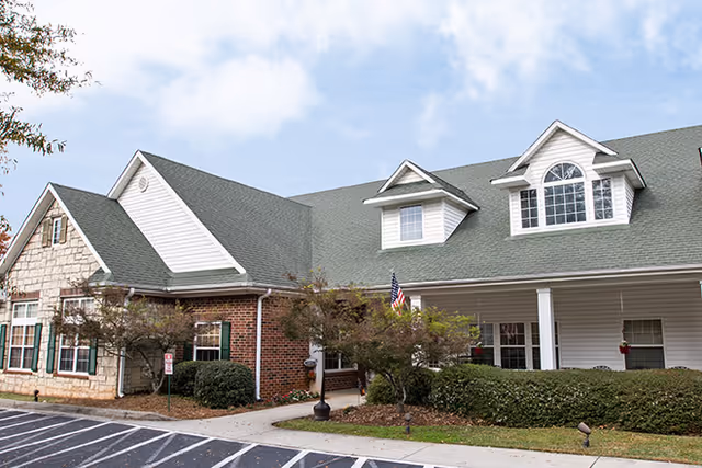 Exterior view of a senior living facility building with a combination of brick, stone, and white siding. The building has a green roof with dormer windows and an American flag near the entrance. There are shrubs and small trees around the entrance and a parking area in front.