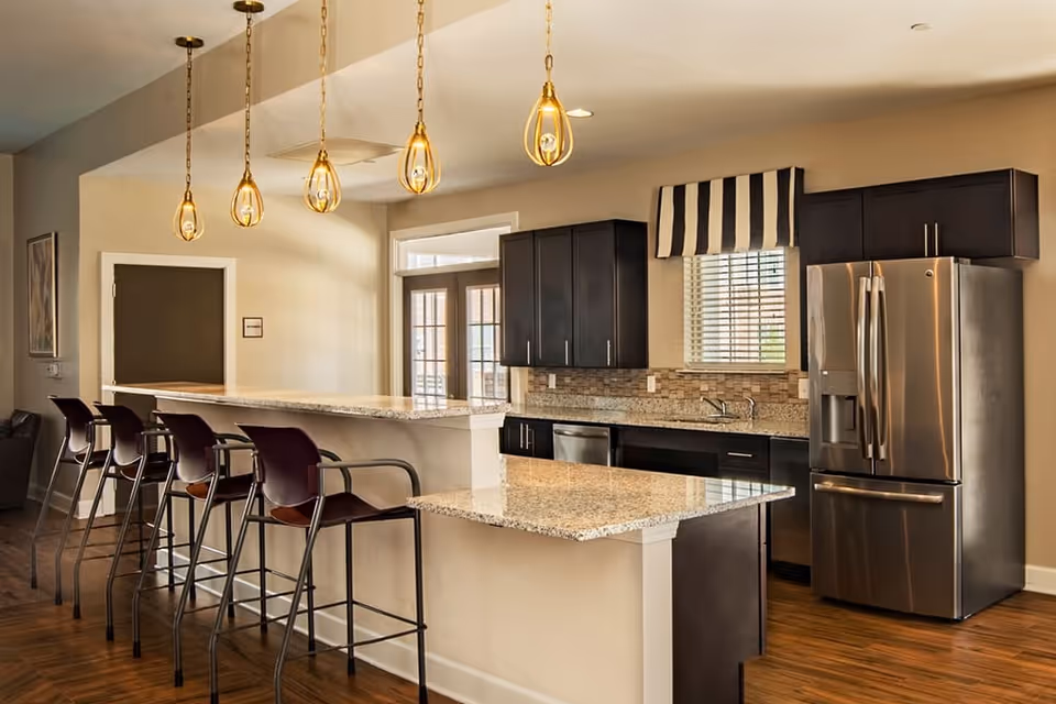Modern kitchen area with granite countertops, dark wood cabinets, stainless steel refrigerator, dishwasher, and sink. Four high chairs are lined up along a breakfast bar under hanging pendant lights. A window with striped valance is above the sink.