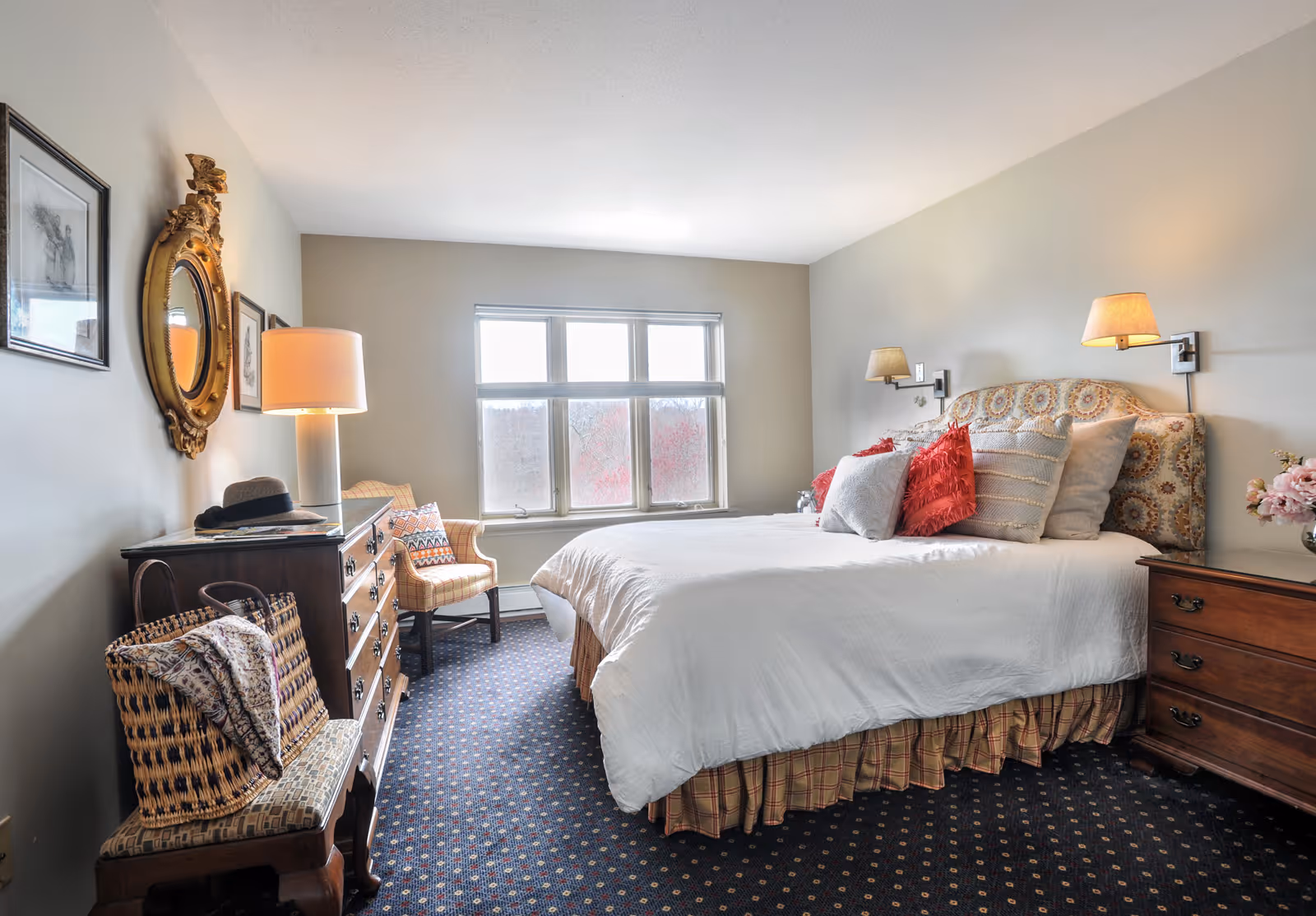 A bright, cozy bedroom with a made bed, patterned headboard, nightstands, dresser, chair, and a window letting in light.