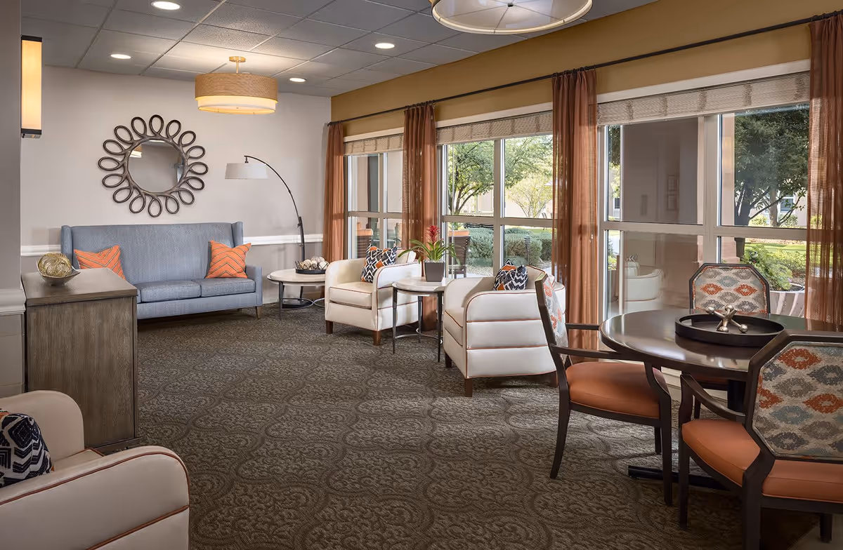A cozy senior living common area with a blue sofa, two white armchairs, a round wooden table with four chairs, and large windows with sheer brown curtains letting in natural light. The room features a decorative wall mirror, a floor lamp, and a patterned carpet.