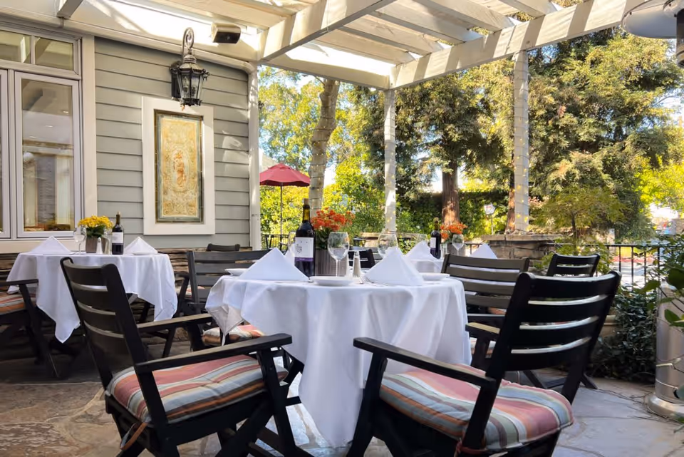 Outdoor patio dining area with tables covered in white tablecloths, set with wine bottles, glasses, and napkins. The space is shaded by a pergola and surrounded by greenery and trees, with cushioned chairs around the tables.
