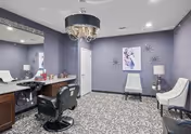 A beauty salon room with two black salon chairs in front of large mirrors and countertops. The walls are painted purple, and there is a patterned carpet on the floor. The room features a chandelier hanging from the ceiling and two white chairs with a small table and lamp between them against the far wall. Decorative wall art is also visible.