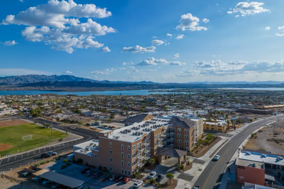 Aerial view of The Views at Lake Havasu Independent Living facility surrounded by roads, parking areas, and nearby residential neighborhoods with a large body of water and mountains in the background under a partly cloudy sky.