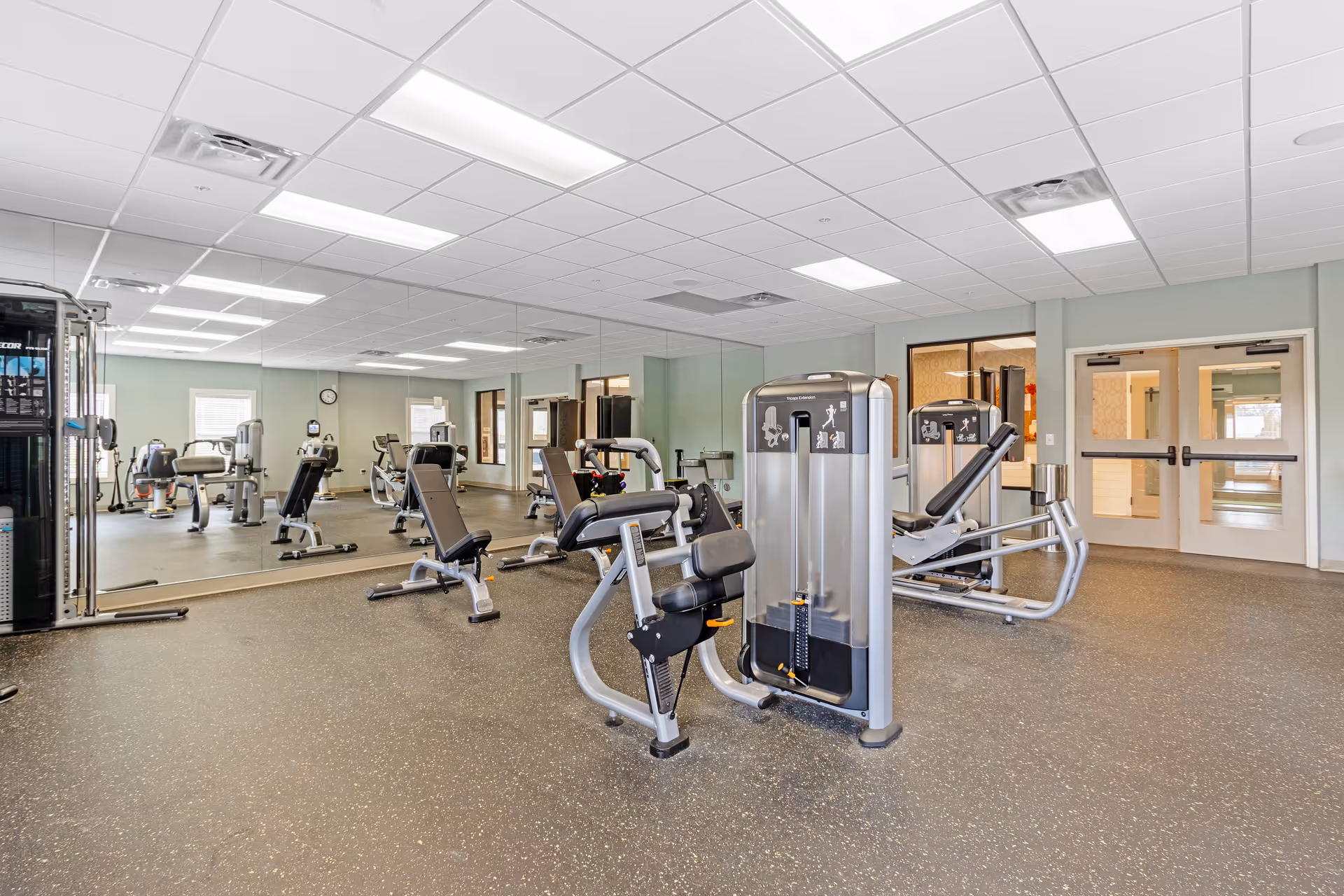 A spacious fitness room with various exercise machines including weight and cardio equipment, large wall mirrors, and bright overhead lighting. The room has a speckled rubber floor and light-colored walls with windows and double doors in the background.