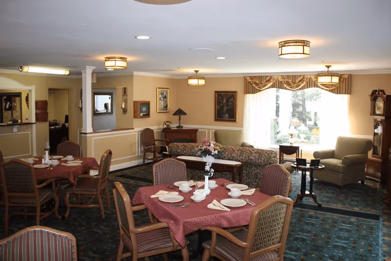 A cozy dining and sitting area in a senior living facility with tables covered in red tablecloths set with plates, bowls, and utensils. There are upholstered chairs around the tables, a floral-patterned sofa, armchairs, a grandfather clock, and framed artwork on beige walls. Large windows with curtains let in natural light, and ceiling lights illuminate the room.