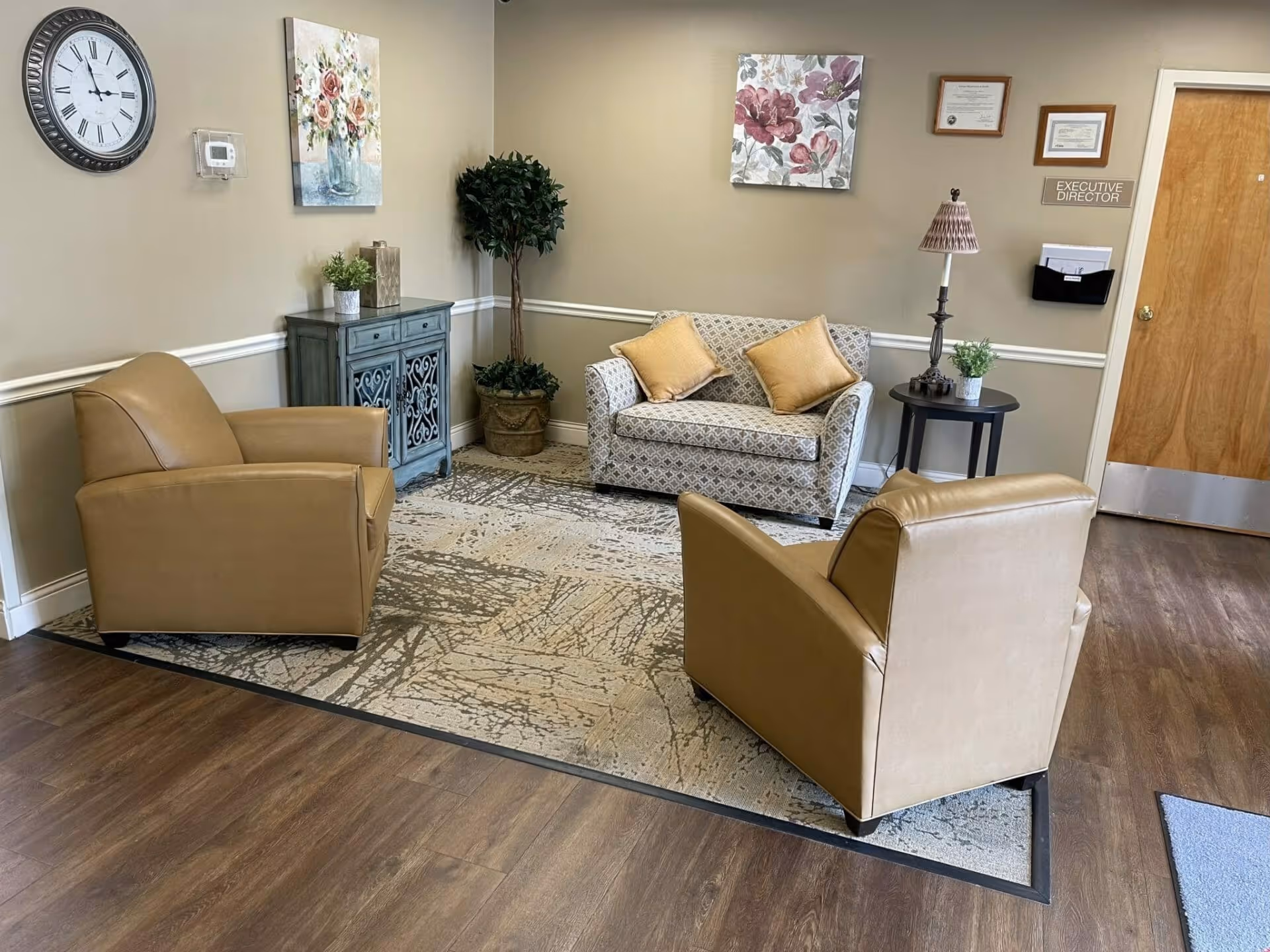 A cozy sitting area with two tan armchairs, a patterned loveseat with throw pillows, a side table and decorative cabinet against a beige wall with artwork and a clock.