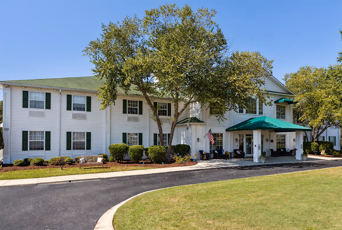 White, two-story senior living facility with green shutters, a covered entrance, and landscaped grounds.