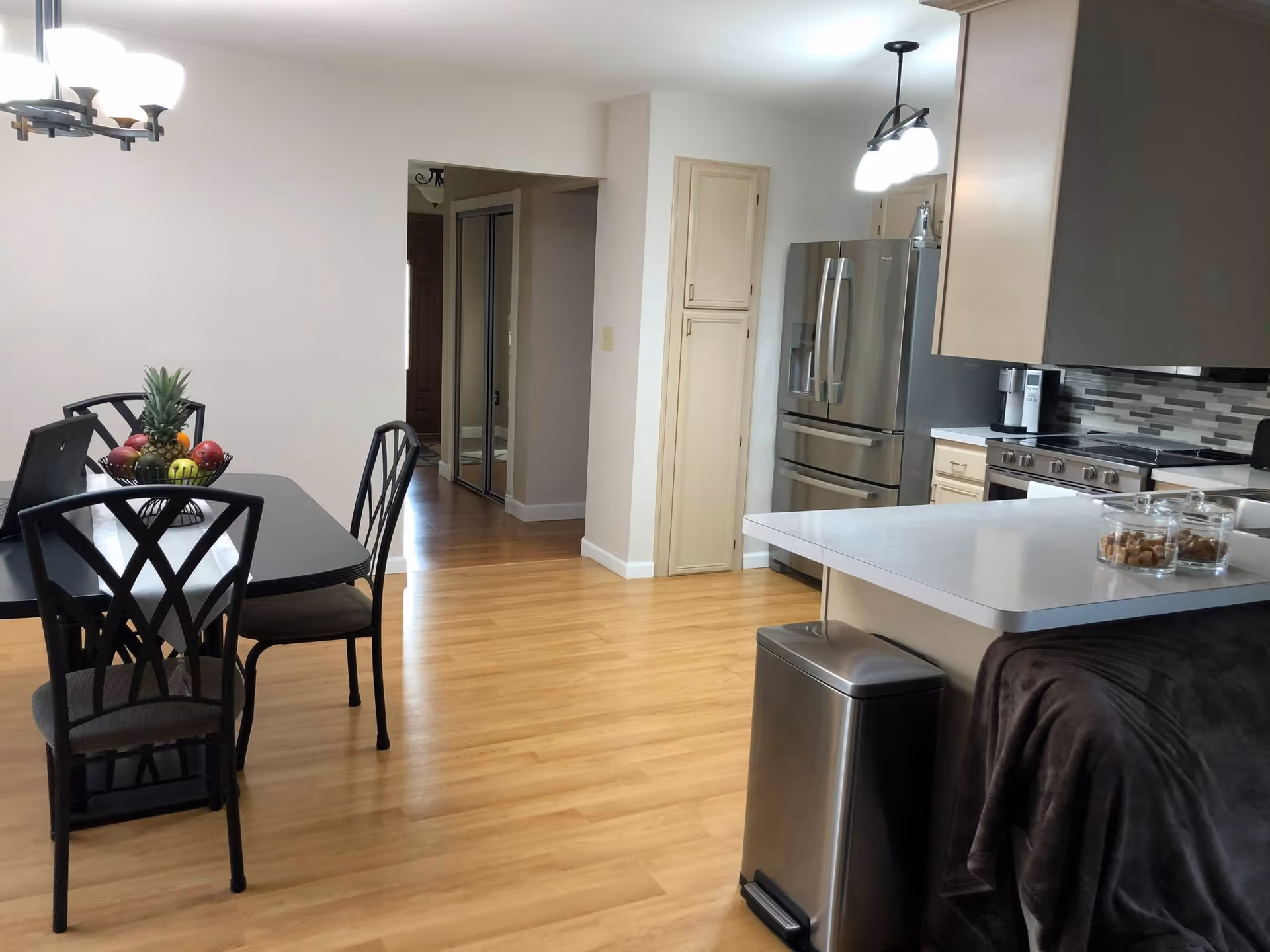 Open kitchen and dining area with a black dining table and chairs, a stainless steel refrigerator and island countertop on hardwood floors.