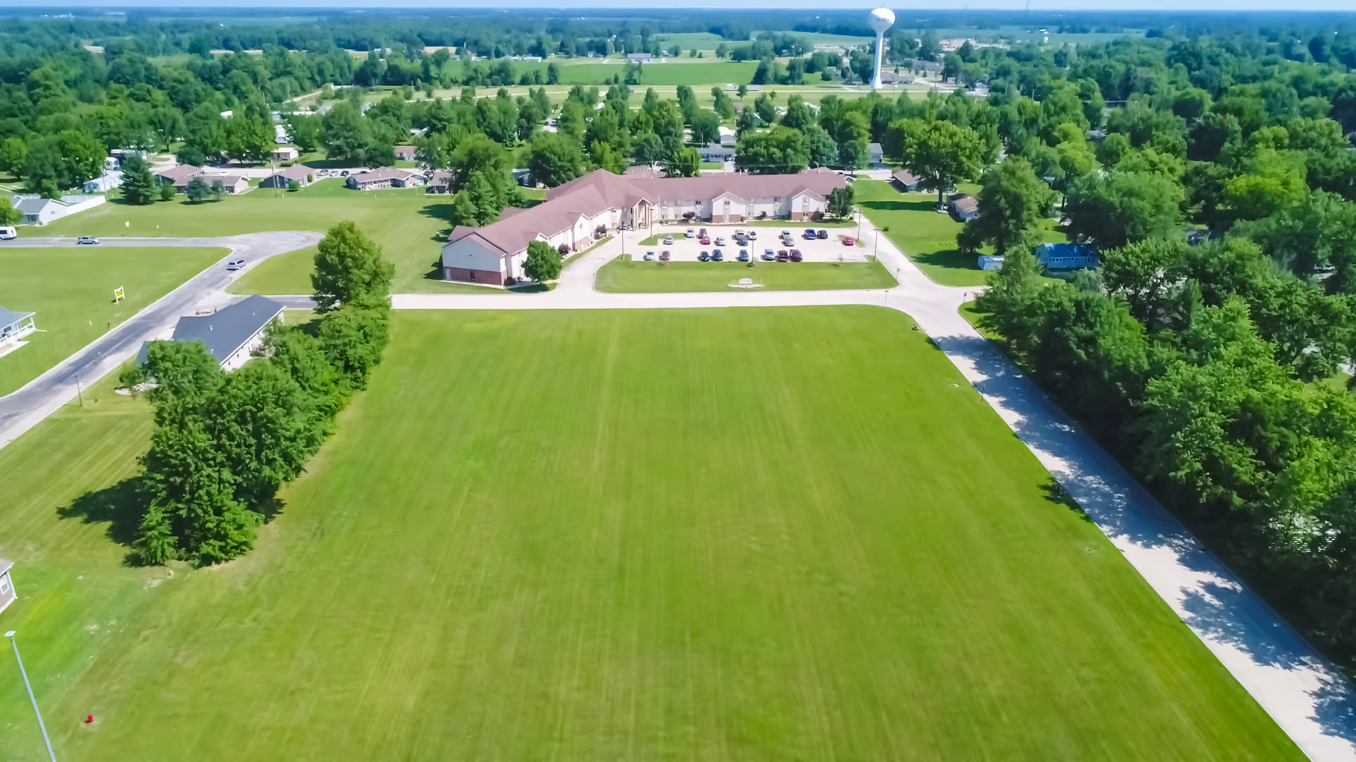 Aerial view of a senior living facility named Heritage Woods of Flora, showing a large green lawn in the foreground, a parking lot with cars, and a long building surrounded by trees and roads under a clear blue sky.
