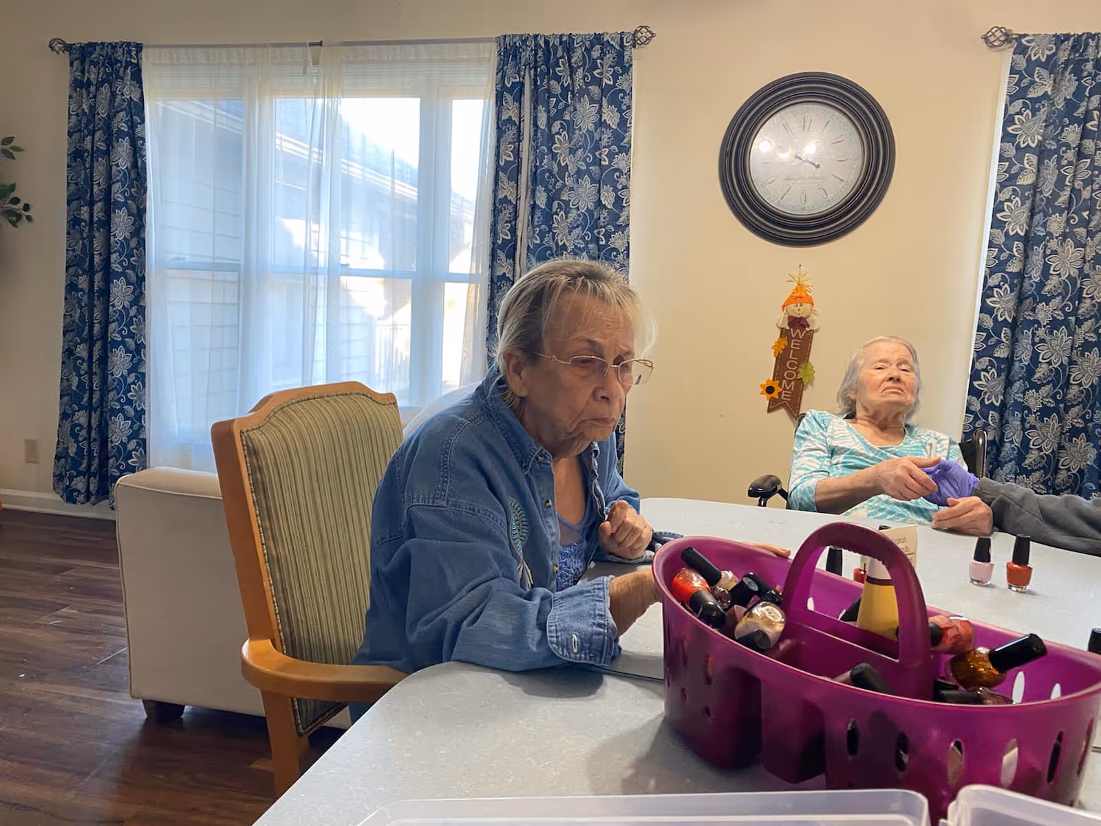 Two elderly women sitting at a table in a room with large windows covered by blue floral curtains. One woman is focused on her hands while the other is having her nails painted. A purple basket filled with various nail polish bottles is on the table. A large clock and a welcome decoration hang on the wall behind them.