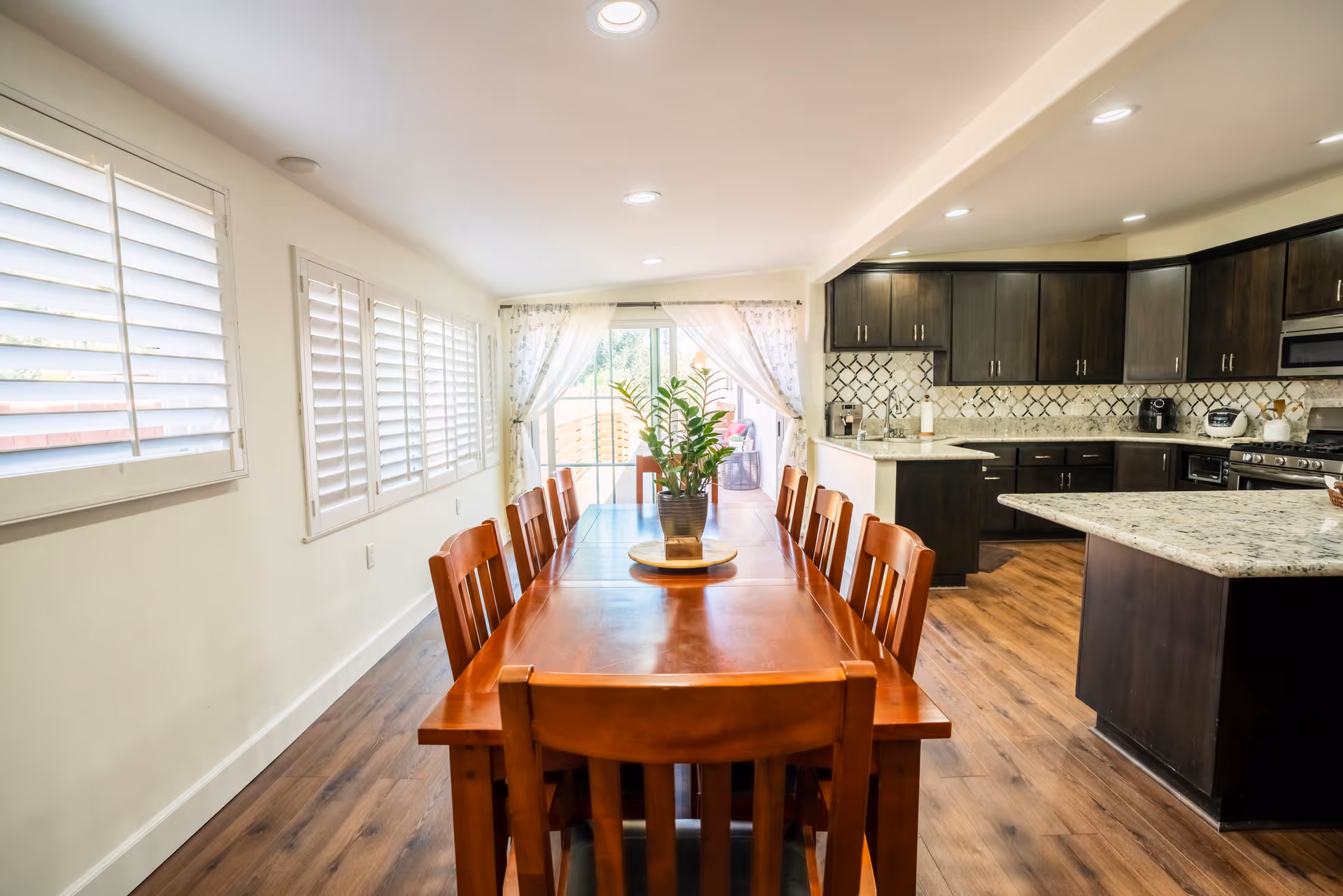 A bright and spacious dining area with a long wooden dining table surrounded by wooden chairs. The table has a small potted plant centerpiece. The room features large windows with white shutters on the left wall and sheer white curtains covering a sliding glass door at the far end, allowing natural light to fill the space. To the right is a modern kitchen with dark wood cabinets, granite countertops, and stainless steel appliances. The floor is a warm-toned wood.