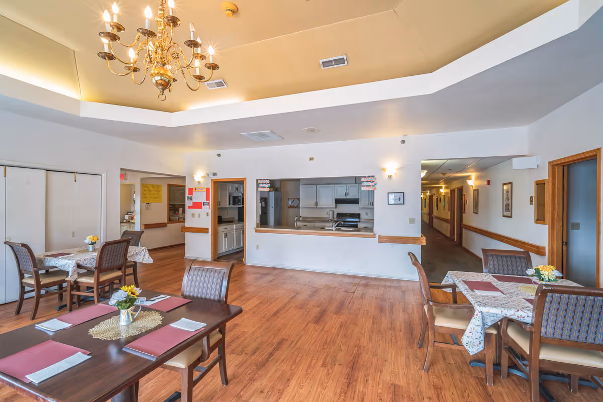 A dining room in a senior living facility with wooden floors, several tables covered with tablecloths and set with placemats and napkins, and a chandelier hanging from the ceiling. The room has a pass-through window to a kitchen area with white cabinets and appliances. There is a hallway visible to the right with framed pictures on the walls.
