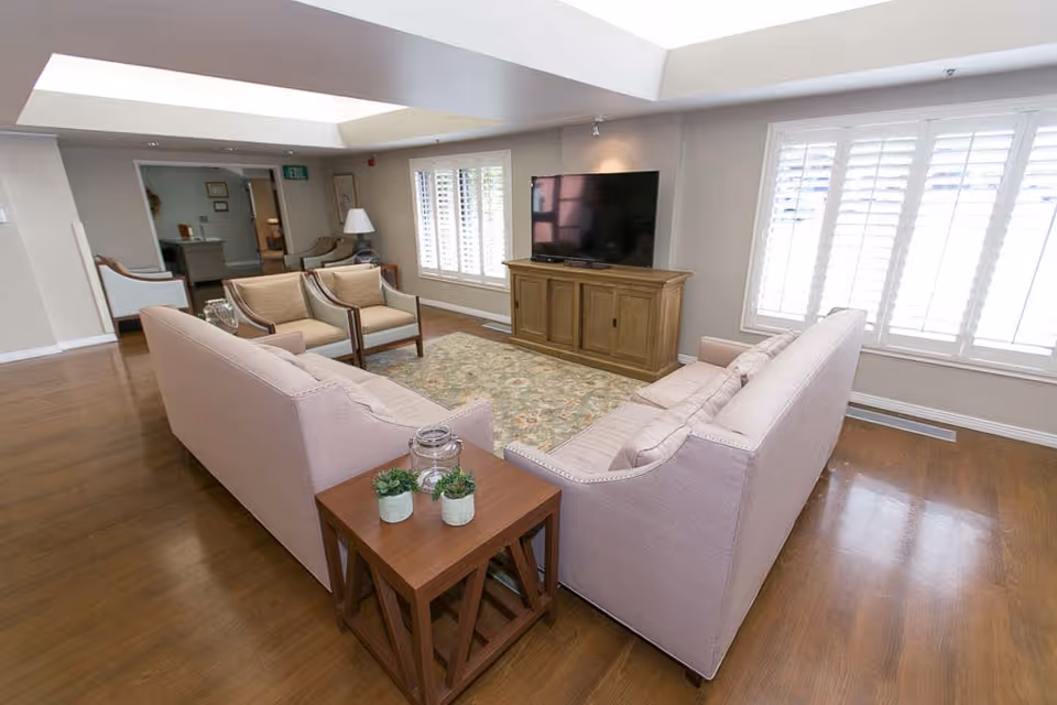 A bright and spacious living room in a senior living facility featuring two beige sofas facing each other with a wooden side table holding small plants and a glass jar. There are two armchairs near a window with white shutters, a large flat-screen TV on a wooden cabinet, and a patterned area rug on the wooden floor.