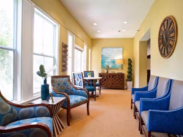 Bright senior living lounge with upholstered armchairs, small tables, artwork, and a large wall clock.