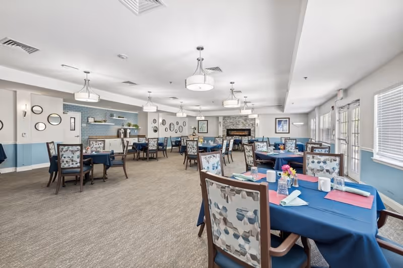 A spacious dining room with multiple tables covered in blue tablecloths, each set with napkins, glasses, and small flower arrangements. The room features patterned chairs, large windows with blinds, modern ceiling lights, and a stone fireplace at the far end. The walls are decorated with round mirrors and framed pictures.