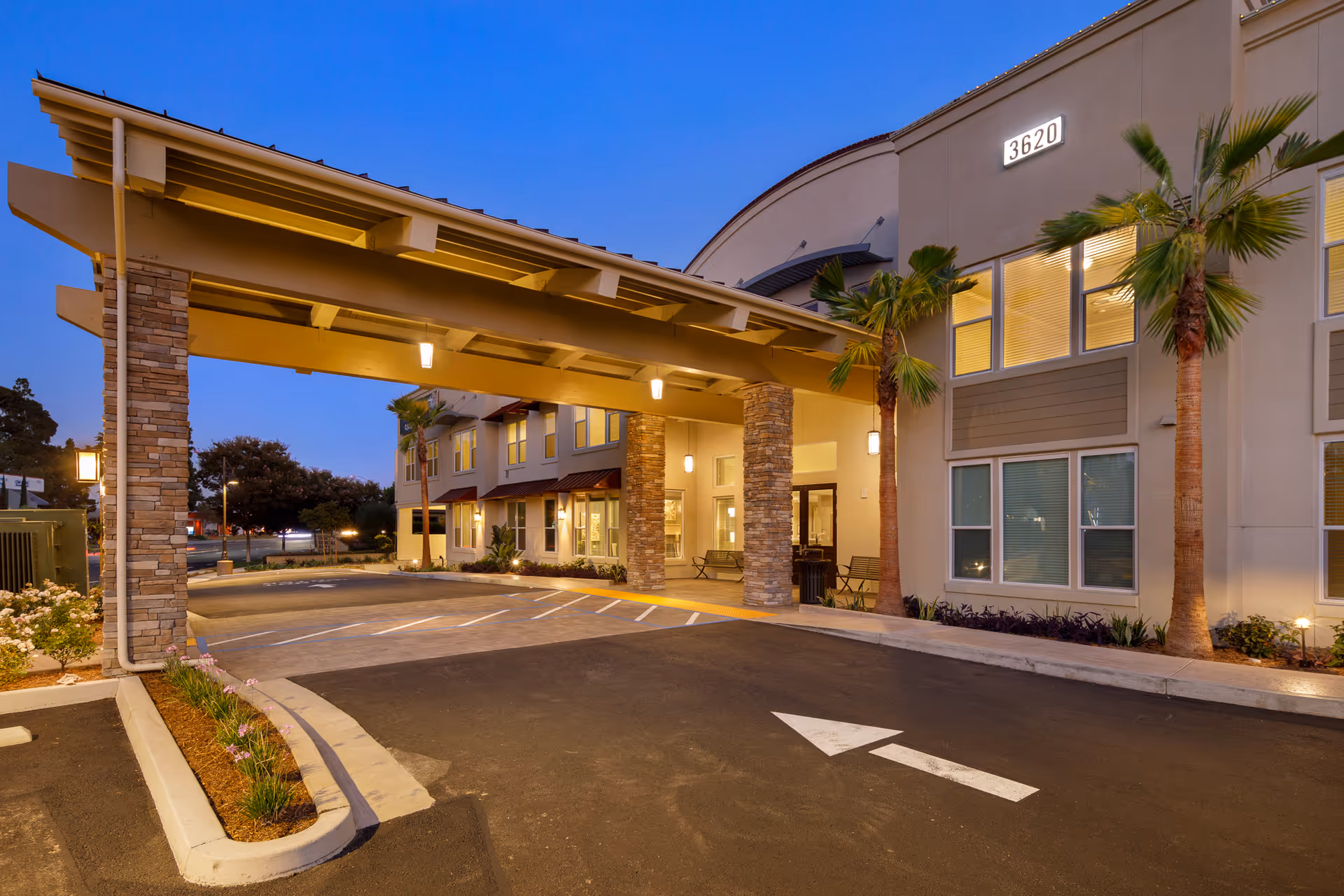 Covered porte-cochere entrance of a senior living building with palm trees, illuminated windows, and a marked driveway at dusk.