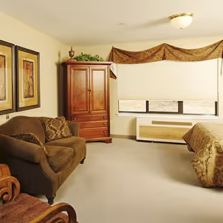 Furnished bedroom with a bed, upholstered sofa, wooden armoire, framed wall art, and a window with shades.