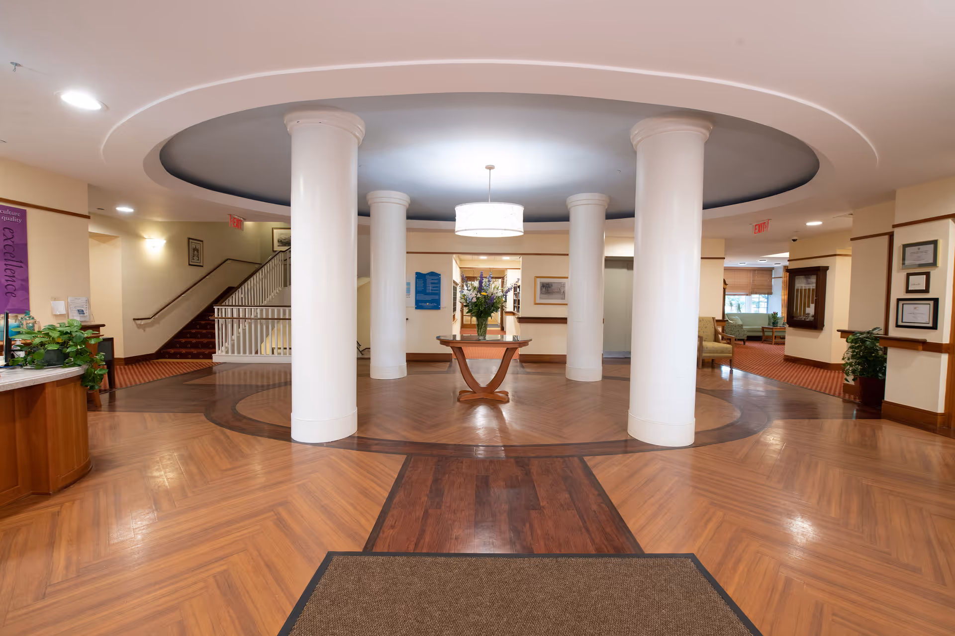 Interior view of a senior living facility lobby with polished wooden floors, four large white columns arranged in a circle, a round ceiling feature with a hanging light fixture, and a small table with a vase of flowers in the center. There are stairs and seating areas visible in the background along with framed pictures and plants.