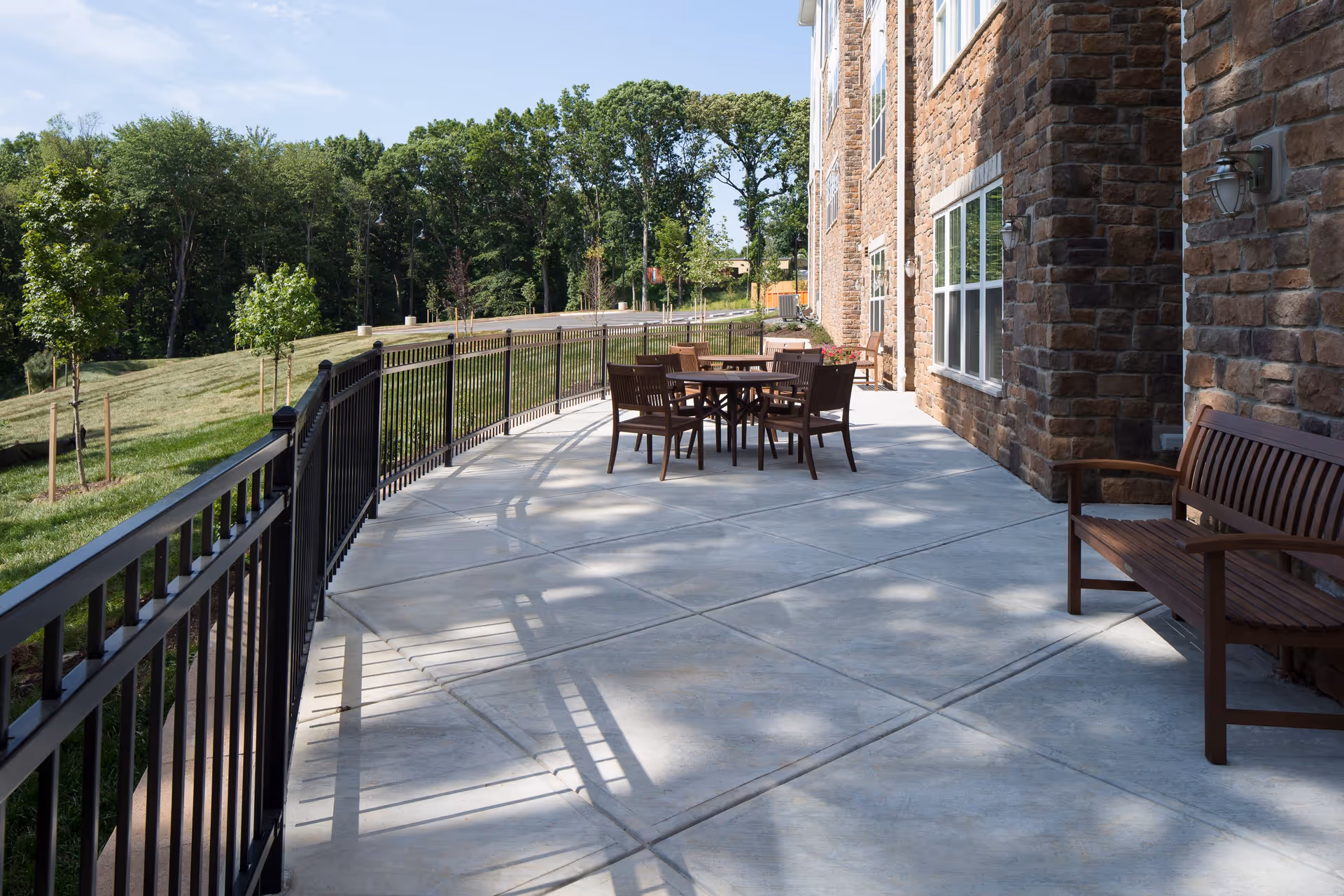 Outdoor patio area at Greens at English Consul with a concrete floor, black metal railing, wooden bench, and a table with chairs. The patio is adjacent to a stone building and overlooks a grassy area with trees.