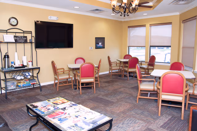 Bright communal dining room with multiple tables and red-upholstered chairs, a wall-mounted TV, coffee station, and a magazine-covered coffee table.