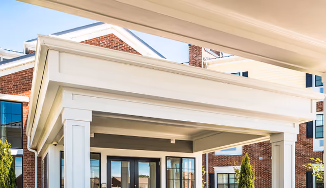 Entrance of a senior living facility with a covered porch supported by white columns, brick exterior walls, and large windows reflecting the sky.
