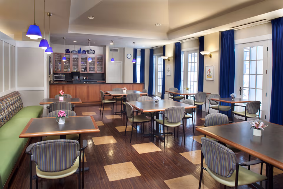 A bright dining room with multiple tables and chairs arranged neatly. The room features a long green cushioned bench along one wall, blue curtains on large windows, and several small flower arrangements on the tables. Blue pendant lights hang from the ceiling above a wooden counter with glass cabinets filled with glassware and decorative items.