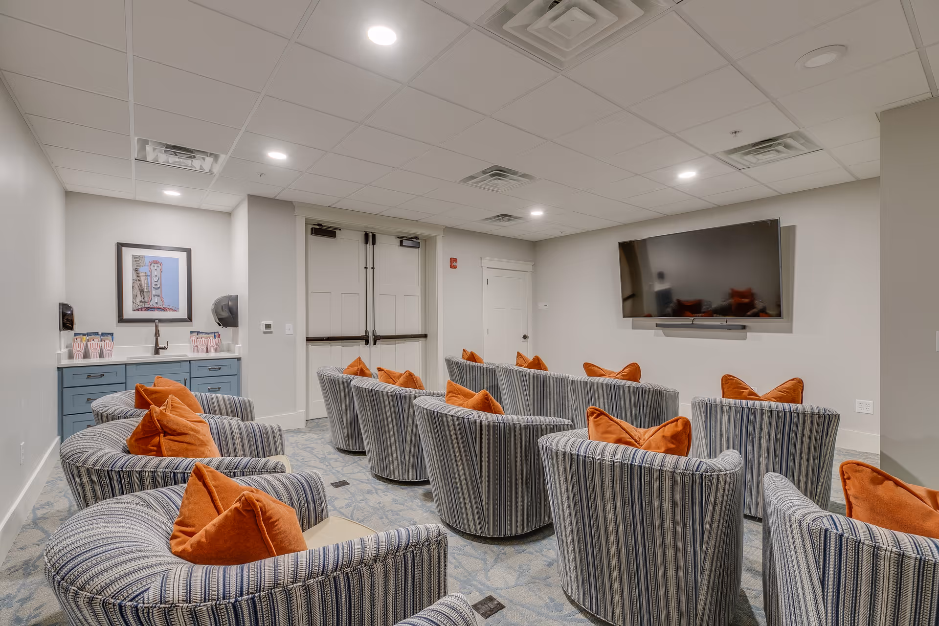 A small theater-style room with multiple striped armchairs each with an orange pillow, facing a large flat-screen TV mounted on the wall. There is a blue cabinet with a sink and popcorn containers on the left side of the room, and double doors at the back.