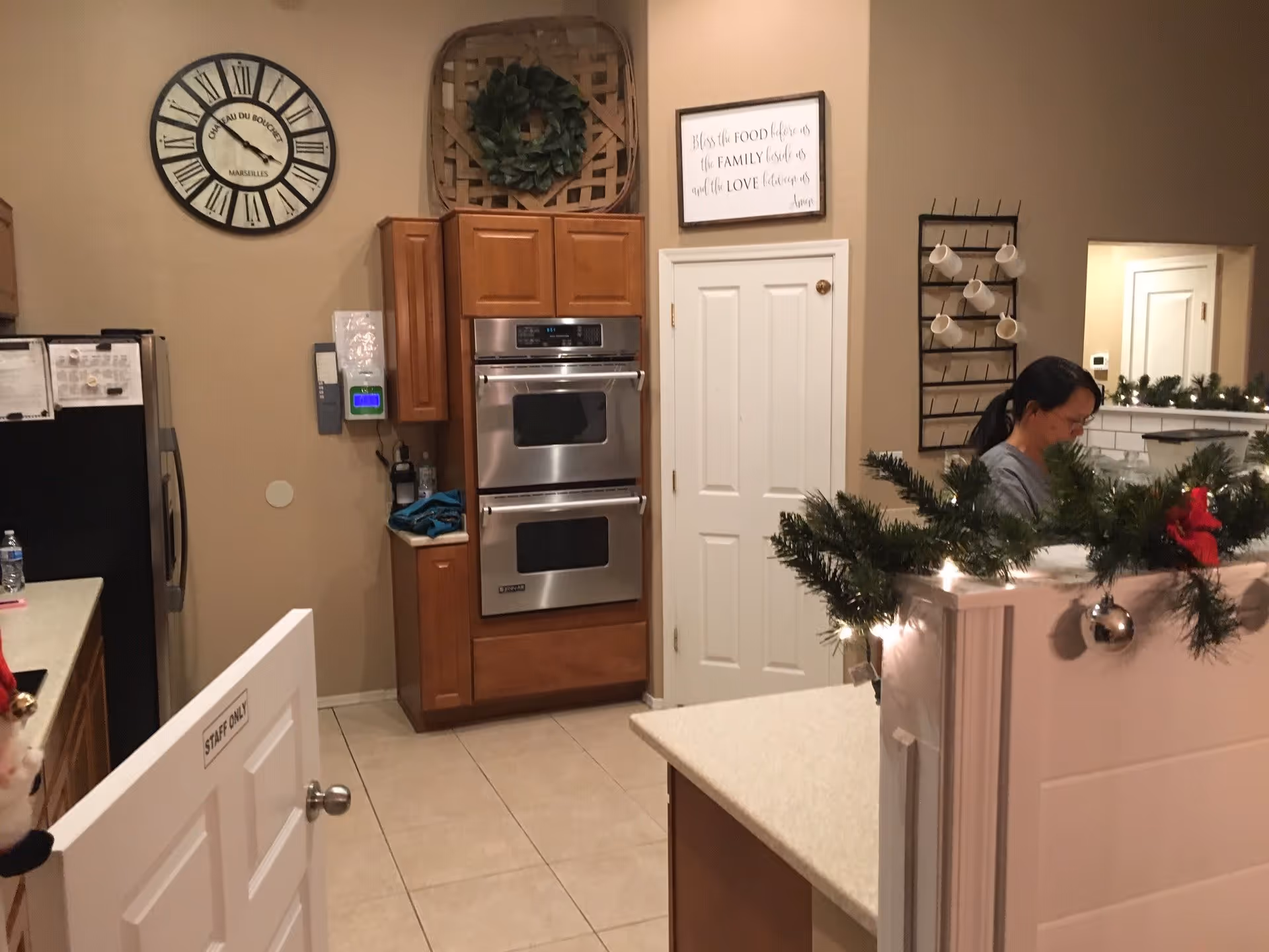 Interior view of a kitchen area with wooden cabinets, double stainless steel ovens, a large wall clock, and a sign that reads 'Bless the food before us the family beside us and the love between us Amen'. A woman is standing near a counter decorated with garland and Christmas lights. A white door with a 'Staff Only' sign is partially open.