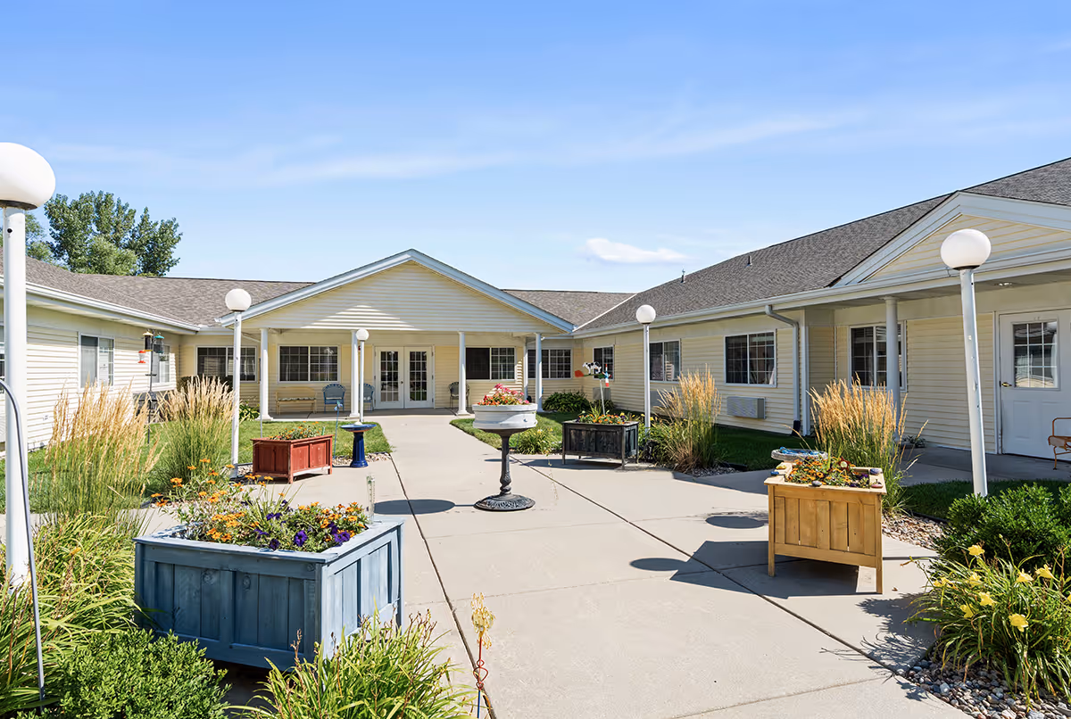 Outdoor courtyard area of Carter Place Senior Living with a concrete walkway, several planters filled with flowers, ornamental grasses, and a few benches. The building surrounding the courtyard is light yellow with white trim and multiple windows. The sky is clear and blue.