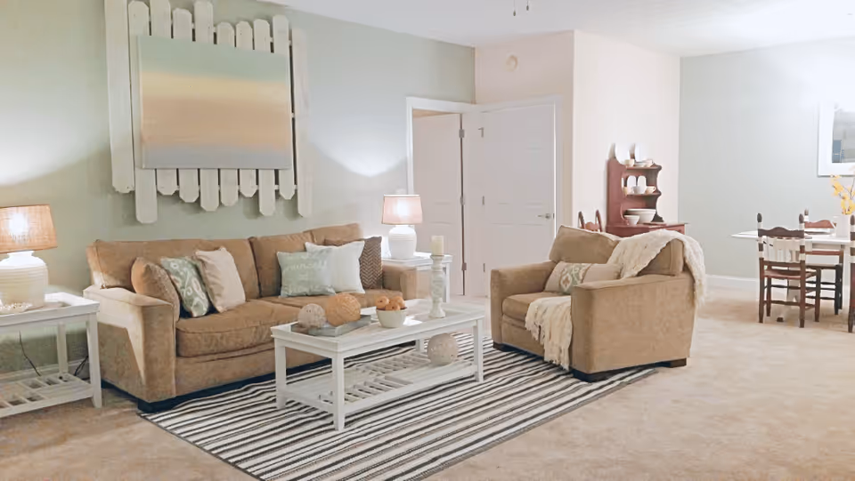 A cozy living room with a beige sofa and armchair, both adorned with decorative pillows and throws. A white coffee table with decorative items sits on a striped rug. Two table lamps with beige shades provide warm lighting. In the background, there is a dining area with a wooden table and chairs, and a small hutch with dishes. The walls are painted in soft neutral tones, and a large abstract painting hangs above the sofa.