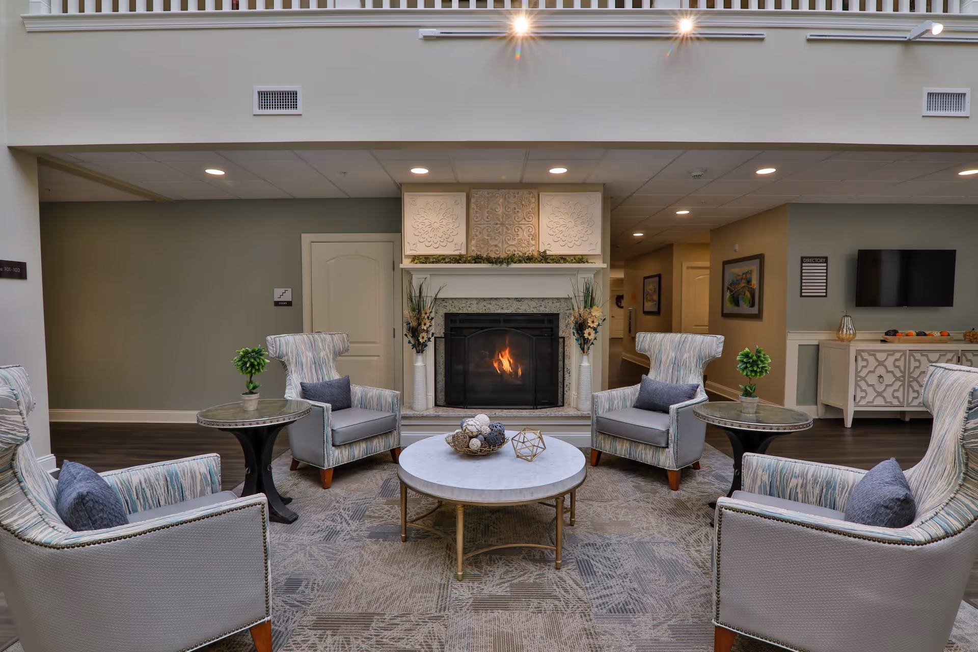 A cozy senior living facility common area with a lit fireplace in the center, surrounded by four upholstered armchairs with blue cushions. There are two round side tables with small potted plants, a round coffee table with decorative items, and a patterned rug on the floor. The walls are painted in neutral tones, and there is a TV mounted on the wall to the right along with a decorative cabinet below it.