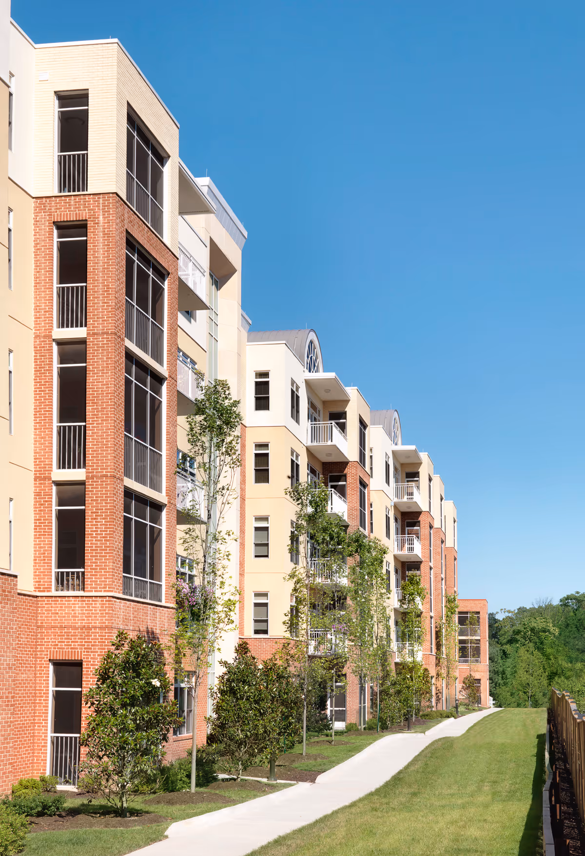Exterior view of a multi-story residential building with a combination of brick and light-colored walls, balconies, and large windows. There is a paved walkway alongside a grassy area with young trees and shrubs under a clear blue sky.