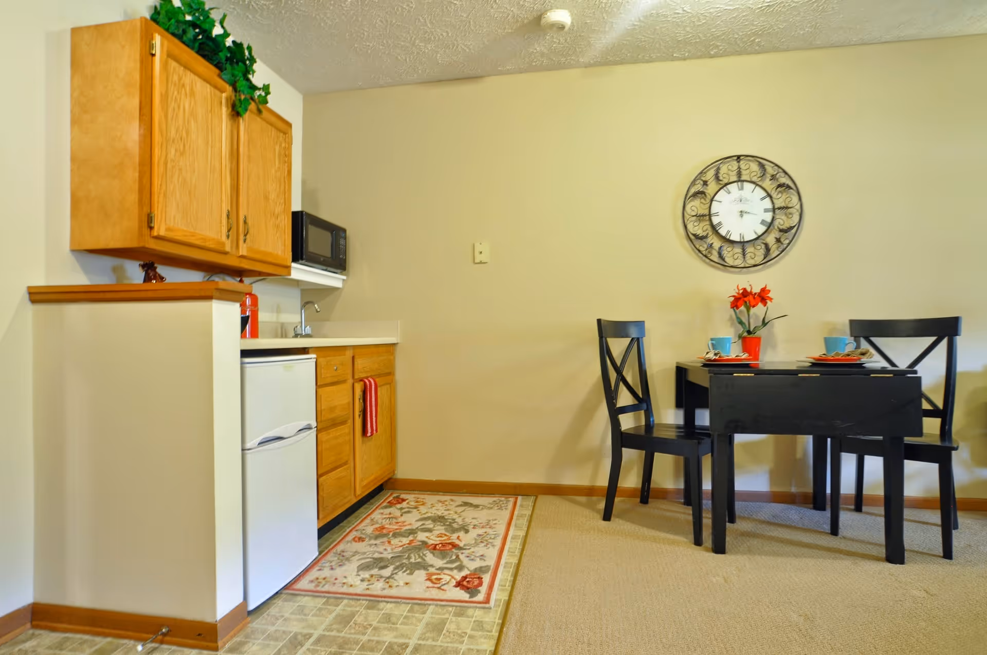 Small kitchenette with wooden cabinets, a mini-fridge and microwave next to a small black dining table with two chairs and a wall clock.
