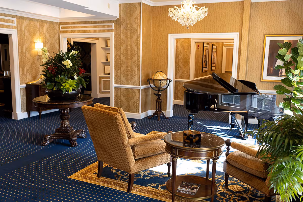 Elegant common room with a black grand piano, upholstered armchairs around a small table, and decorative plants and lighting.