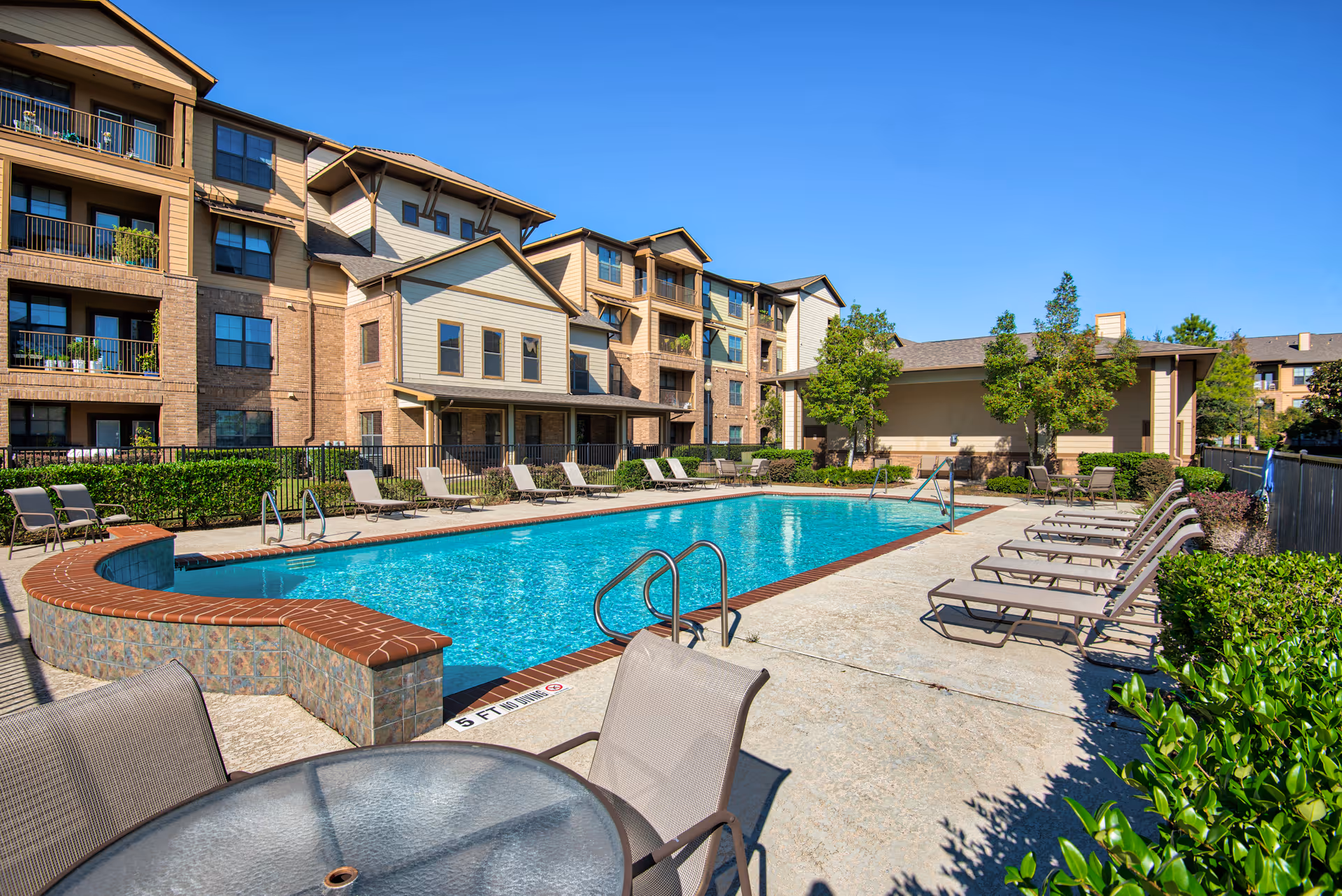 Outdoor swimming pool area at a senior living facility with lounge chairs arranged around the pool, a round glass table with chairs in the foreground, and multi-story residential buildings in the background under a clear blue sky.