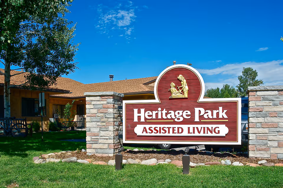 A large red and white 'Heritage Park Assisted Living' sign mounted between stone pillars in front of a single-story building with grass and trees.