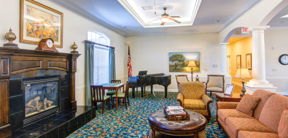 A cozy living room area in a senior living facility featuring a fireplace with a wooden mantle, a framed painting above it, a small table with chairs near a window with curtains, a grand piano, an armchair, a sofa, a round leather ottoman with magazines, a side table with a lamp, and a ceiling fan with lights. The room has patterned carpet and light-colored walls with decorative molding and columns.