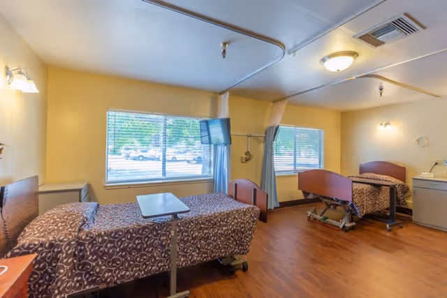 A nursing facility room with two hospital beds covered in patterned bedspreads, each equipped with an overbed table. The room has large windows with blinds, a wall-mounted TV, and light yellow walls. The floor is wood, and there are privacy curtains between the beds.