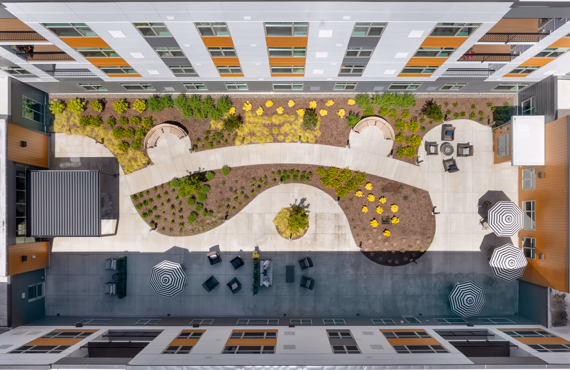 Aerial view of an outdoor courtyard at The Trinity Community at Miami Township featuring a curved concrete walkway, landscaped garden beds with various plants and shrubs, circular seating areas, patio chairs arranged around a fire pit, and several tables with striped umbrellas.