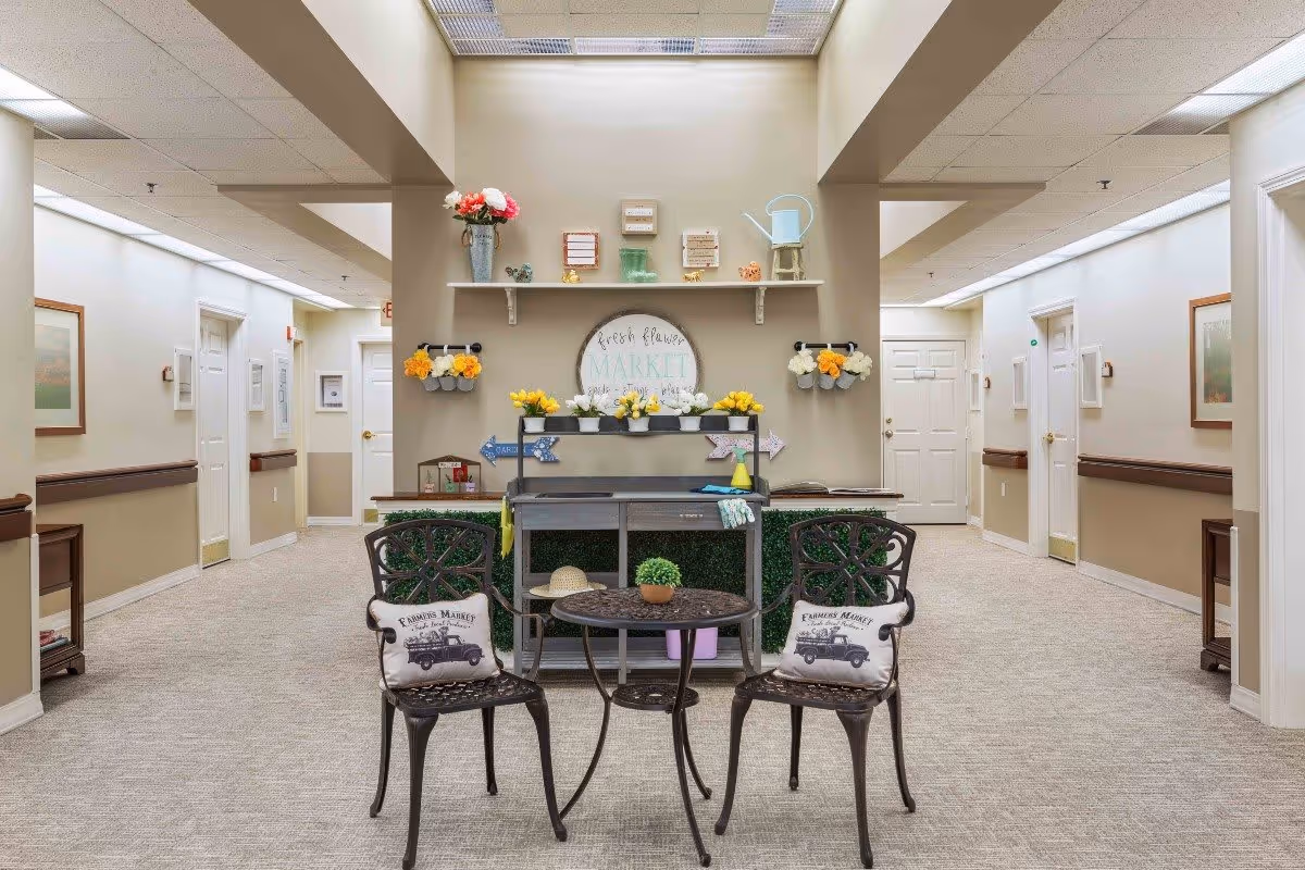 A bright hallway in a senior living facility with beige walls and carpeted floors. In the center, there is a small decorative seating area with two black metal chairs featuring cushions that say 'Farmers Market' and a round metal table with a small plant. Behind the seating area is a decorative shelf with various flower pots, a watering can, and a sign that reads 'Fresh Flower Market'. The hallway has several doors and framed pictures on the walls.