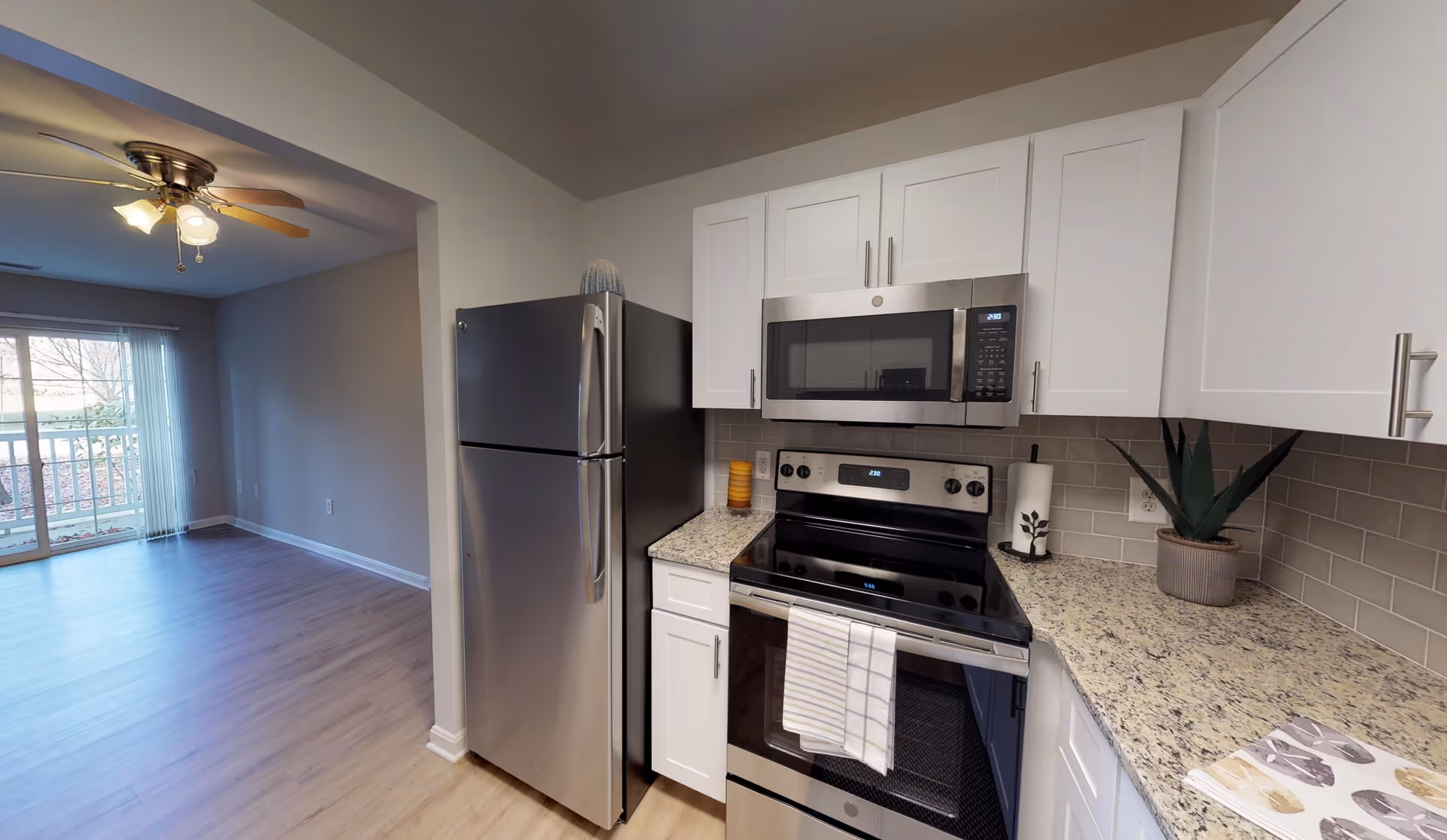 Modern kitchen area with stainless steel refrigerator, stove, and microwave. White cabinets and granite countertops with a small potted plant and paper towel holder. Adjacent room with ceiling fan and sliding glass door leading to a balcony or outdoor space.