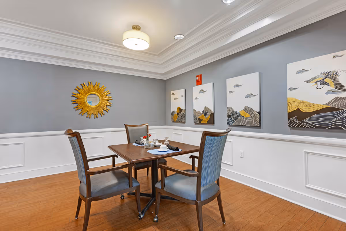 A small dining area with a wooden table and four cushioned chairs on wheels. The table is set with a white cup and saucer, a small vase with flowers, and some napkins. The room has light gray walls with white wainscoting, wooden flooring, and a ceiling light fixture. On the walls are decorative paintings featuring birds and abstract landscapes, and a sunburst mirror.