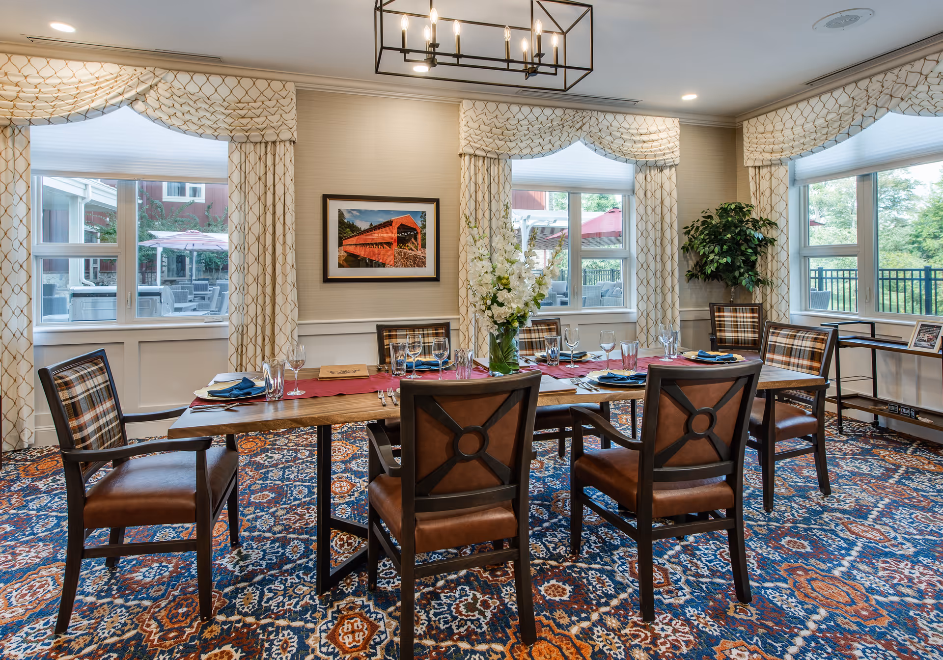 A dining room with a wooden table set for six people with plates, glasses, and napkins. The room has large windows with patterned curtains, a colorful patterned carpet, a chandelier overhead, a framed picture on the wall, and a potted plant in the corner.