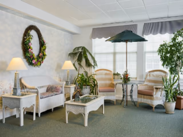 Sunlit sitting room with white wicker sofa and chairs, a glass-top coffee table, potted plants, lamps and a wreath on the wall.