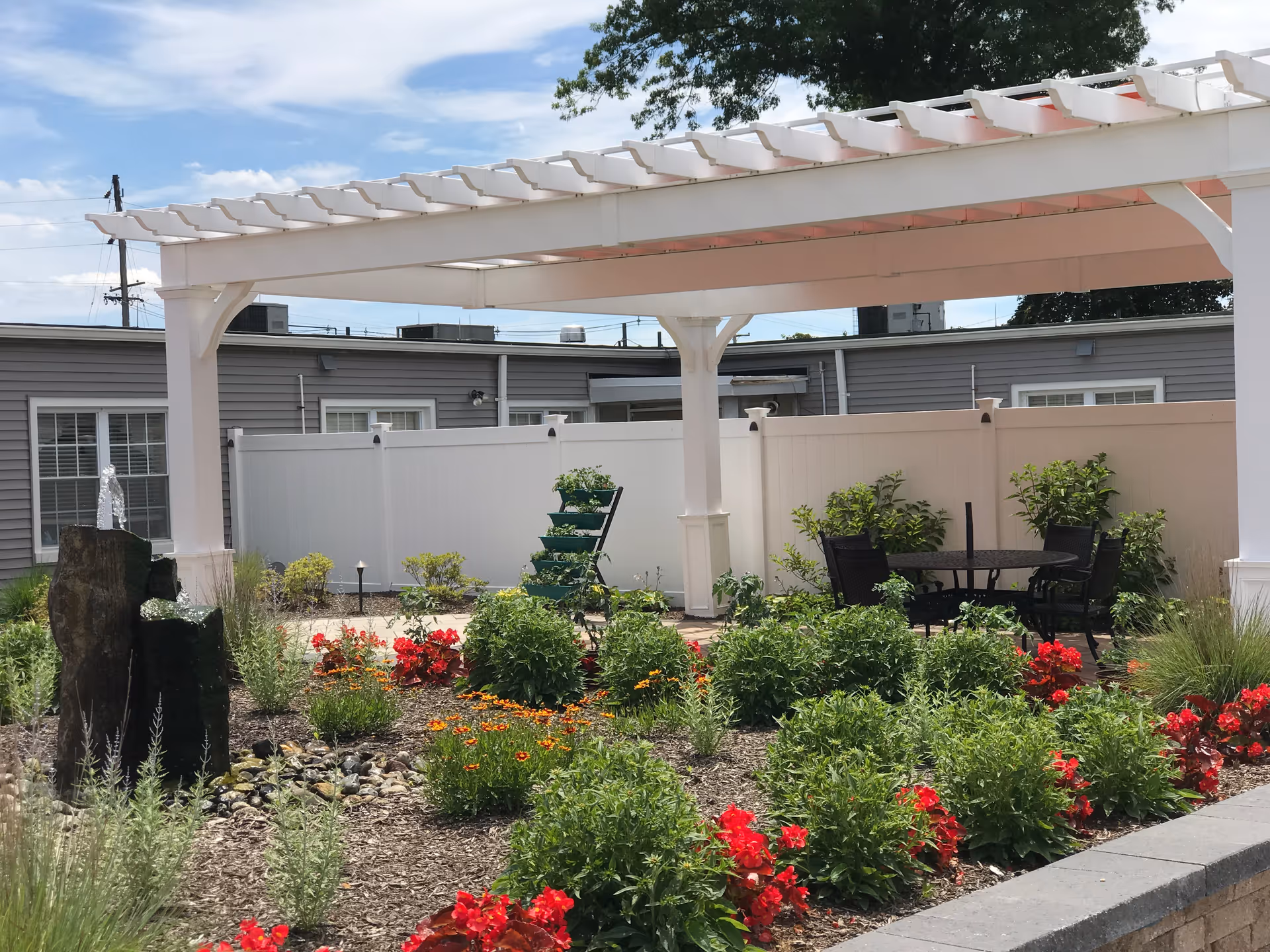Outdoor garden area with a white pergola, green shrubs, red and orange flowers, a small water fountain feature made of dark stone, and a black metal table with chairs under the pergola. The background shows a gray building with white windows and a white fence.