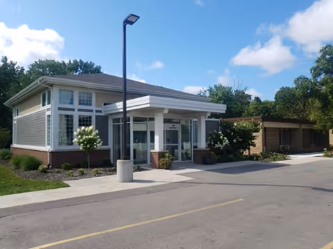 Exterior view of a single-story building with large windows and a covered entrance, surrounded by greenery and a paved driveway under a partly cloudy sky.