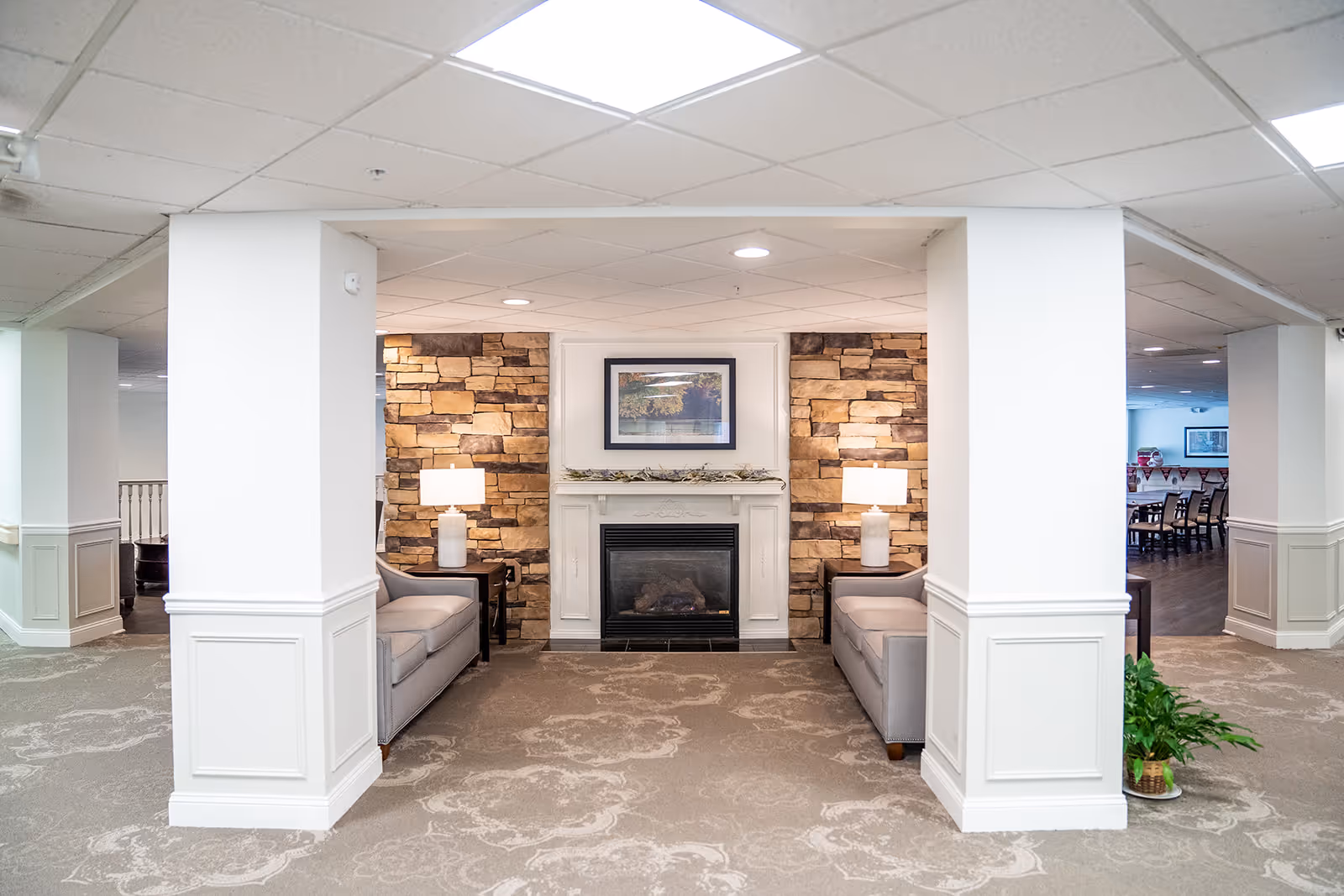 A cozy seating area in a senior living facility featuring two gray sofas facing each other with a white fireplace in the center. The fireplace is flanked by stone accent walls and two table lamps on dark side tables. Above the fireplace is a framed landscape painting. The space is carpeted with a patterned design, and white pillars frame the seating area. In the background, a dining area with tables and chairs is visible.