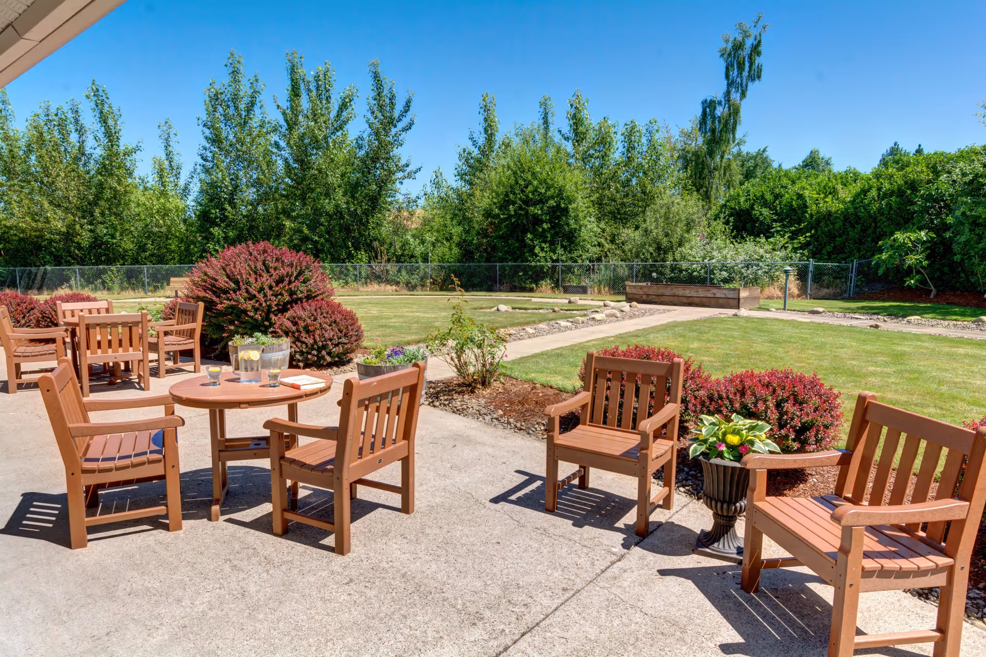 Outdoor patio area with wooden chairs and tables on a concrete surface, surrounded by green grass, bushes, and trees under a clear blue sky.