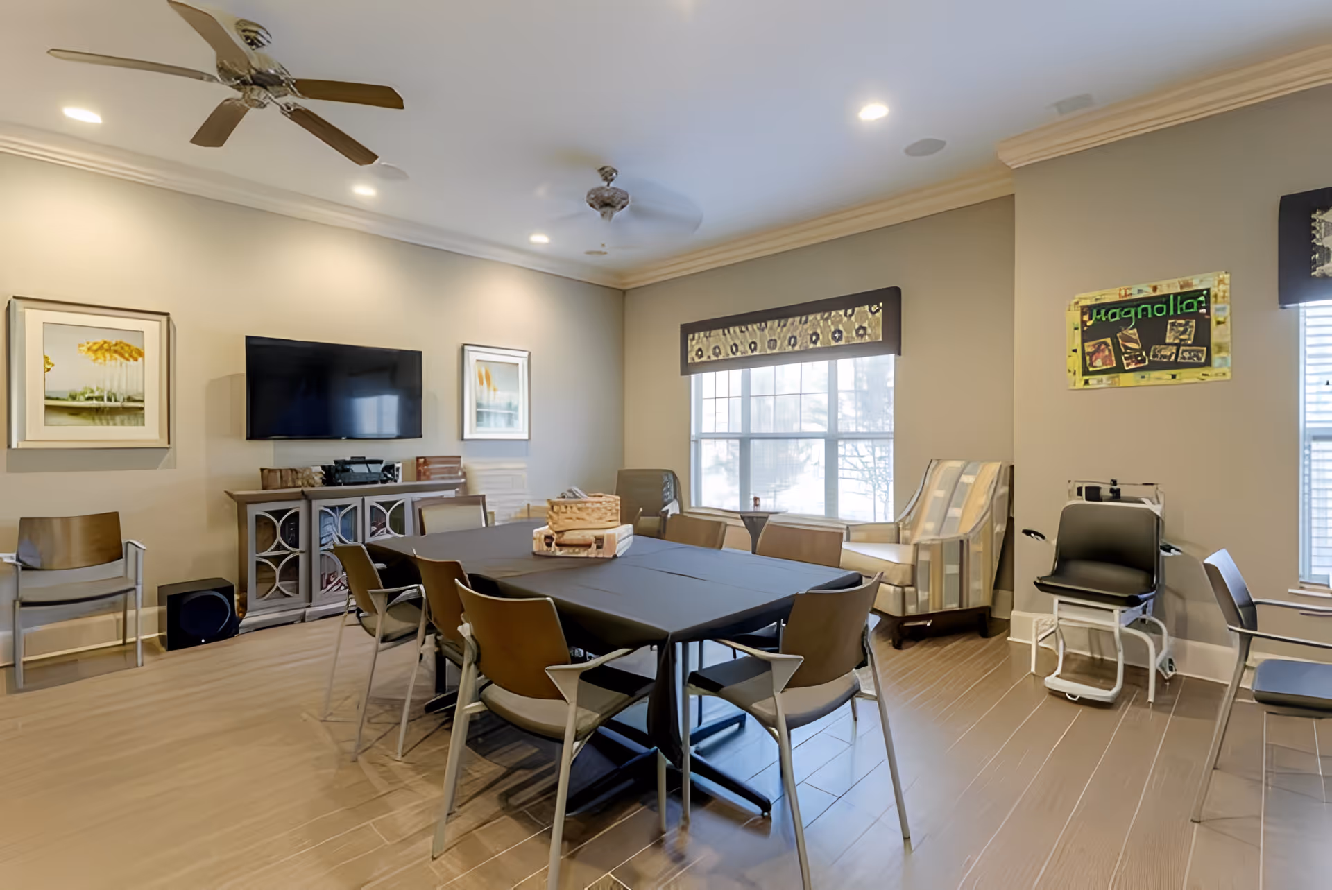 A well-lit common room with a rectangular table covered with a black tablecloth surrounded by chairs. The room features two ceiling fans, a wall-mounted flat-screen TV, framed artwork on the walls, large windows with patterned valances, and additional seating including armchairs. The floor is wooden, and the walls are painted a light beige color.